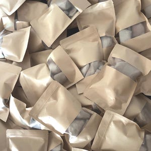 May include: A large collection of light brown kraft paper pouches with clear plastic windows. Each pouch is sealed at the top and contains small, irregular white objects visible through the transparent section of the packaging.