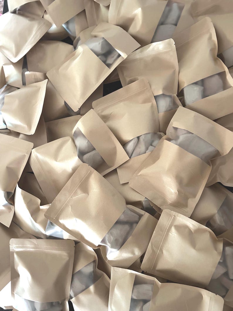May include: A large collection of light brown kraft paper pouches with clear plastic windows. Each pouch is sealed at the top and contains small, irregular white objects visible through the transparent section of the packaging.