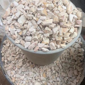 May include: A large, round, grey plastic bucket filled to the brim with small, jagged, light beige and off-white stone fragments. The bucket is overflowing with these irregular, crushed rocks, which are also scattered around the base of the container.