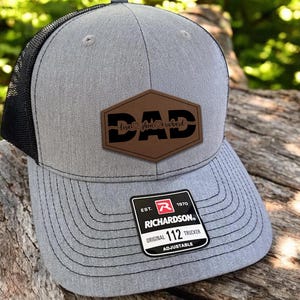 May include: Gray trucker hat with a black mesh back. The hat features a brown leather patch with the word "DAD" and the names "Lisa, Linda, Richard". The hat has a curved brim and an adjustable strap.