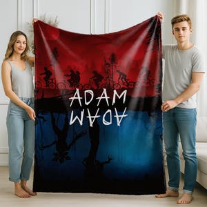 K&ouml;nnte beinhalten: Eine rot-blaue Decke mit einer Silhouette von Radfahrern vor rotem Himmel. Die Decke tr&auml;gt den wei&szlig;en Schriftzug "ADAM", darunter der gleiche Text gespiegelt. Die untere H&auml;lfte der Decke ist blau mit einer dunklen Baumsilhouette.