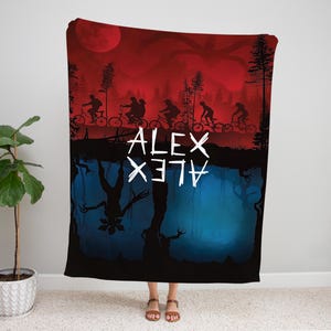 K&ouml;nnte beinhalten: Eine schwarze Decke mit einem rot-blauen Grafikdesign. Die obere H&auml;lfte zeigt Silhouetten von Menschen auf Fahrr&auml;dern unter einem roten Himmel mit einem gro&szlig;en Mond. Die untere H&auml;lfte zeigt eine umgekehrte blaue Landschaft mit einem Monster. Der Name "ALEX" ist wei&szlig; geschrieben.