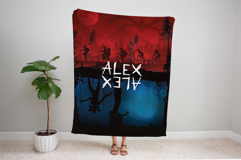K&ouml;nnte beinhalten: Eine schwarze Decke mit einem rot-blauen Grafikdesign. Die obere H&auml;lfte zeigt Silhouetten von Menschen auf Fahrr&auml;dern unter einem roten Himmel mit einem gro&szlig;en Mond. Die untere H&auml;lfte zeigt eine umgekehrte blaue Landschaft mit einem Monster. Der Name "ALEX" ist wei&szlig; geschrieben.