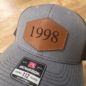 May include: A grey trucker hat featuring a brown leather-like hexagonal patch centered on the front panel with the number 1998 engraved in black. A black rectangular sticker on the brim displays the text Richardson 112 Trucker Est 1970.