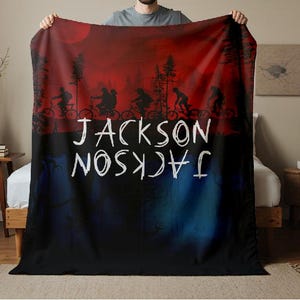 K&ouml;nnte beinhalten: Eine personalisierte Decke mit einer rot-schwarzen Silhouette von Radfahrern unter einem roten Mond. Der Name "JACKSON" ist in Wei&szlig; geschrieben, sowohl richtig als auch verkehrt herum.