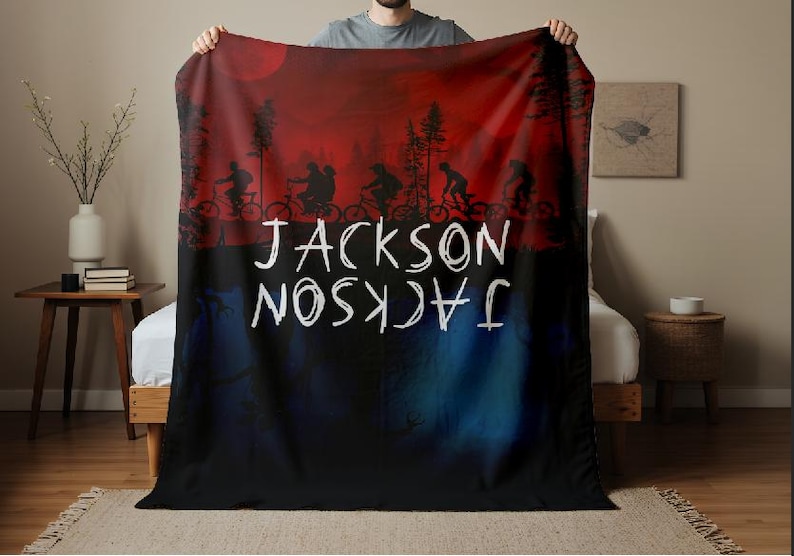 K&ouml;nnte beinhalten: Eine personalisierte Decke mit einer rot-schwarzen Silhouette von Radfahrern unter einem roten Mond. Der Name "JACKSON" ist in Wei&szlig; geschrieben, sowohl richtig als auch verkehrt herum.