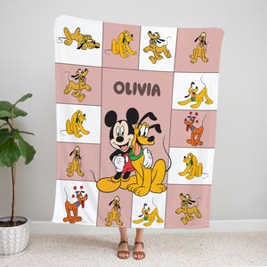 May include: A personalized blanket featuring Mickey Mouse and Pluto, with the name "OLIVIA" in brown letters. The blanket has a pink and white grid pattern with Pluto illustrations in various poses. The blanket is made of soft material.