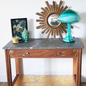 Peut inclure: Une console en bois avec un plateau en marbre et une étagère inférieure dorée à la feuille. La table a un seul tiroir avec une quincaillerie en laiton ornée. Une lampe en verre vert avec un abat-jour blanc et un miroir soleil doré sont sur la table.