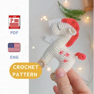 May include: Crochet pattern for a white unicorn toy with red mane and tail, embroidered with stars. The unicorn has wooden wheels and is held in a hand.