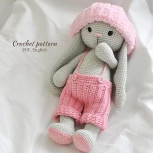 Puede incluir: Una muñeca de conejito hecha a mano con ganchillo, con un mono rosa y un sombrero a juego. El conejito es gris con detalles en rosa en los pies y el mono. El texto "Crochet pattern PDF, English" es visible.