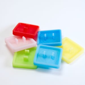 May include: A collection of colorful, square-shaped outlet covers in red, blue, pink, green, and yellow. Each cover has a raised center and two small prongs for insertion into an electrical outlet. The covers are made of a translucent plastic material.
