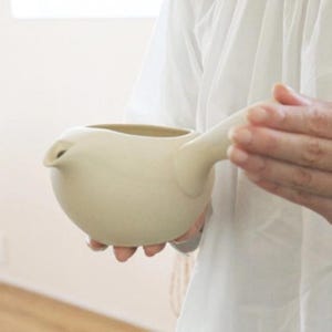 May include: A cream-colored ceramic pitcher with a rounded body and a long, curved handle. The pitcher has a spout and is held by a person wearing a white shirt. The design is minimalist and modern.