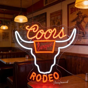 May include: Neon sign in the shape of a bull's head with the words "Coors Banquet" and "Rodeo" in orange and white neon. The sign is mounted on a clear backing and is in a restaurant setting.