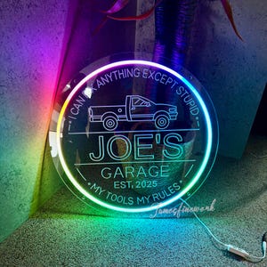 May include: A round, illuminated sign with the text "I CAN FIX ANYTHING EXCEPT STUPID" and "JOE'S GARAGE EST. 2025." A line drawing of a pickup truck is in the center. The sign is lit with a rainbow gradient.