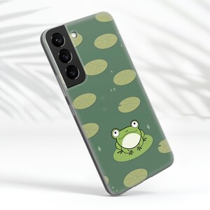 K&ouml;nnte beinhalten: Eine gr&uuml;ne Handyh&uuml;lle mit einem Frosch- und Seerosen-Design. Die H&uuml;lle hat eine transparente R&uuml;ckseite und einen gr&uuml;nen Rand. Das Kameraobjektiv ist auf der R&uuml;ckseite der H&uuml;lle sichtbar.