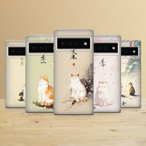 Könnte beinhalten: Fünf Handyhüllen mit verschiedenen Designs, die Katzen und japanische Schriftzeichen zeigen. Die Hüllen sind alle weiß mit durchsichtiger Rückseite und haben einen schwarzen Kamerabuckel. Die Designs beinhalten eine weiße Katze mit rotem Halsband, eine braun-weiße Katze, eine weiße Katze mit rosa Halsband und eine weiße Katze mit blauem Halsband. Die japanischen Schriftzeichen auf den Hüllen sind alle unterschiedlich und in schwarzer Tinte geschrieben.