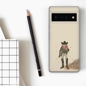 K&ouml;nnte beinhalten: Eine beige Handyh&uuml;lle mit einem gr&uuml;nen Frosch, der einen Cowboyhut und ein rotes Bandana tr&auml;gt. Der Frosch h&auml;lt eine Pistole und hat einen entschlossenen Ausdruck. Der Hintergrund ist hellbraun mit kleinen goldenen Sternen.
