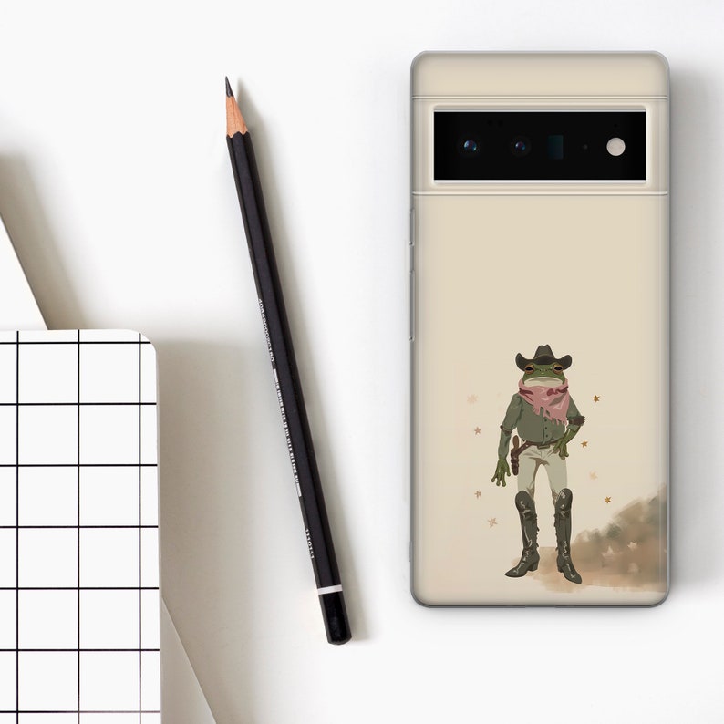 K&ouml;nnte beinhalten: Eine beige Handyh&uuml;lle mit einem gr&uuml;nen Frosch, der einen Cowboyhut und ein rotes Bandana tr&auml;gt. Der Frosch h&auml;lt eine Pistole und hat einen entschlossenen Ausdruck. Der Hintergrund ist hellbraun mit kleinen goldenen Sternen.