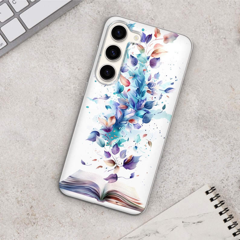 Op de afbeelding: Een witte telefoonhoes met een aquarelontwerp van blauwe en paarse bladeren en een open boek. De telefoonhoes heeft een drievoudige camera lens.