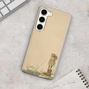 K&ouml;nnte beinhalten: Eine transparente Handyh&uuml;lle mit beigefarbenem Hintergrund und einem Comic-Frosch, der einen rot-wei&szlig;en Pilz-Hut tr&auml;gt. Der Frosch steht auf einer Wiese mit wei&szlig;en Blumen.