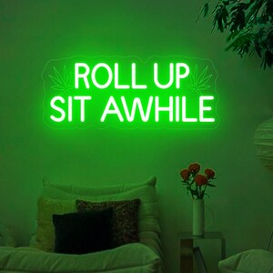 Puede incluir: Letrero de neón verde con el texto "Roll Up Sit Awhile" y una hoja de marihuana estilizada a cada lado del texto.