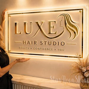 May include: A rectangular wall sign for Luxe Hair Studio featuring gold lettering and a stylised profile of a woman with flowing hair. The sign is backlit with a warm glow and includes the text Beauty, Confidence, You on a clear acrylic panel.