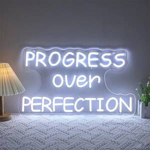 May include: White neon sign with the text "PROGRESS over PERFECTION" on a white background.