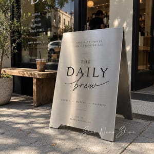 Pode incluir: Uma placa de calçada em forma de A de metal prateado escovado com texto em preto que diz THE DAILY BREW, SPECIALTY COFFEE FOR A BRIGHTER DAY, COFFEE, MATCHA, PASTRIES e EST. 2023 SANTA MONICA, CA. A placa tem um acabamento metálico limpo.