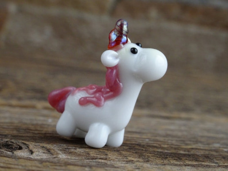Op de afbeelding: Een klein wit glazen eenhoornbeeldje met een roze manen en staart. De eenhoorn heeft een rood en iriserend hoorn.