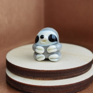 May include: A small glass figurine of a gray sloth with black eyes and a white belly. The sloth is sitting with its arms crossed and has a happy expression on its face.