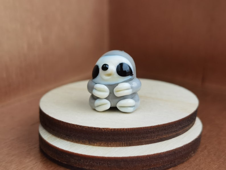 May include: A small glass figurine of a gray sloth with black eyes and a white belly. The sloth is sitting with its arms crossed and has a happy expression on its face.