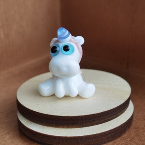 May include: A small white glass unicorn figurine with blue and pink accents. The unicorn has large blue eyes and is sitting on a wooden base.