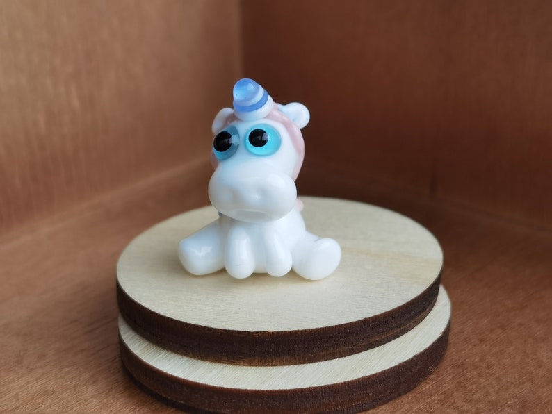 May include: A small white glass unicorn figurine with blue and pink accents. The unicorn has large blue eyes and is sitting on a wooden base.