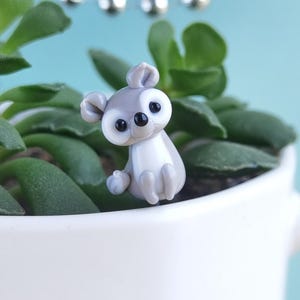 Może przedstawiać: Mała, szaro-biała figurka koali z czarnymi oczami siedzi w białej doniczce wypełnionej zielonymi sukulentami. Koala ma duże uszy i pozuje jakby siedział. W tle sznur srebrnych koralików.
