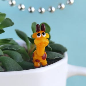 May include: A small, yellow giraffe figurine with brown spots and antlers stands in a white ceramic pot with green plants. A string of silver beads hangs in the background against a light blue backdrop.