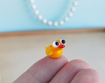 Puede incluir: Una pequeña figurita de pájaro de vidrio amarillo con grandes ojos negros y un pico rojo. El pájaro está sentado en un dedo.