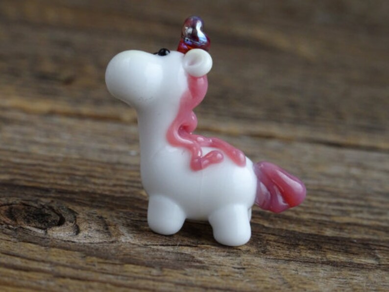 Op de afbeelding: Een klein wit glazen eenhoornbeeldje met roze en paarse accenten. De eenhoorn heeft een roze manen en staart en een paarse hoorn.
