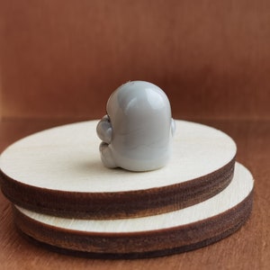 May include: A small, gray, ceramic figurine of a person sitting with their legs crossed. The figurine is sitting on a wooden platform.
