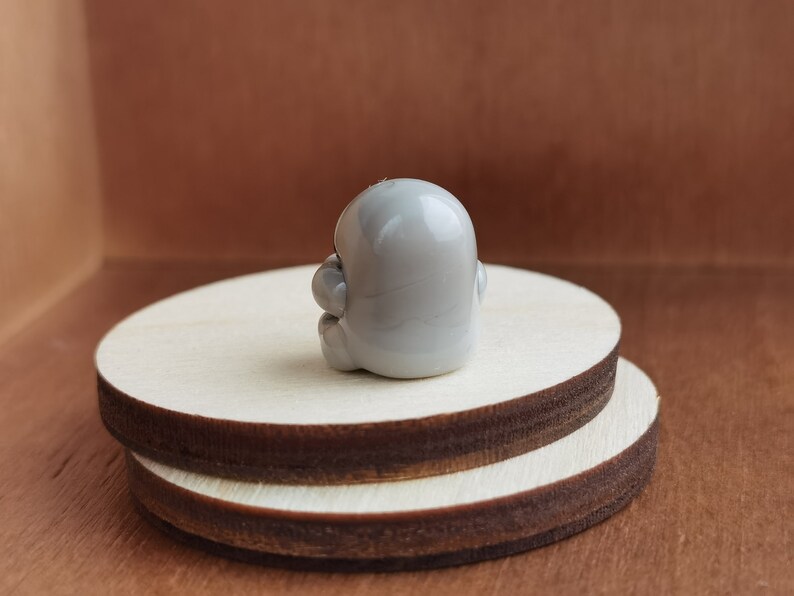 May include: A small, gray, ceramic figurine of a person sitting with their legs crossed. The figurine is sitting on a wooden platform.
