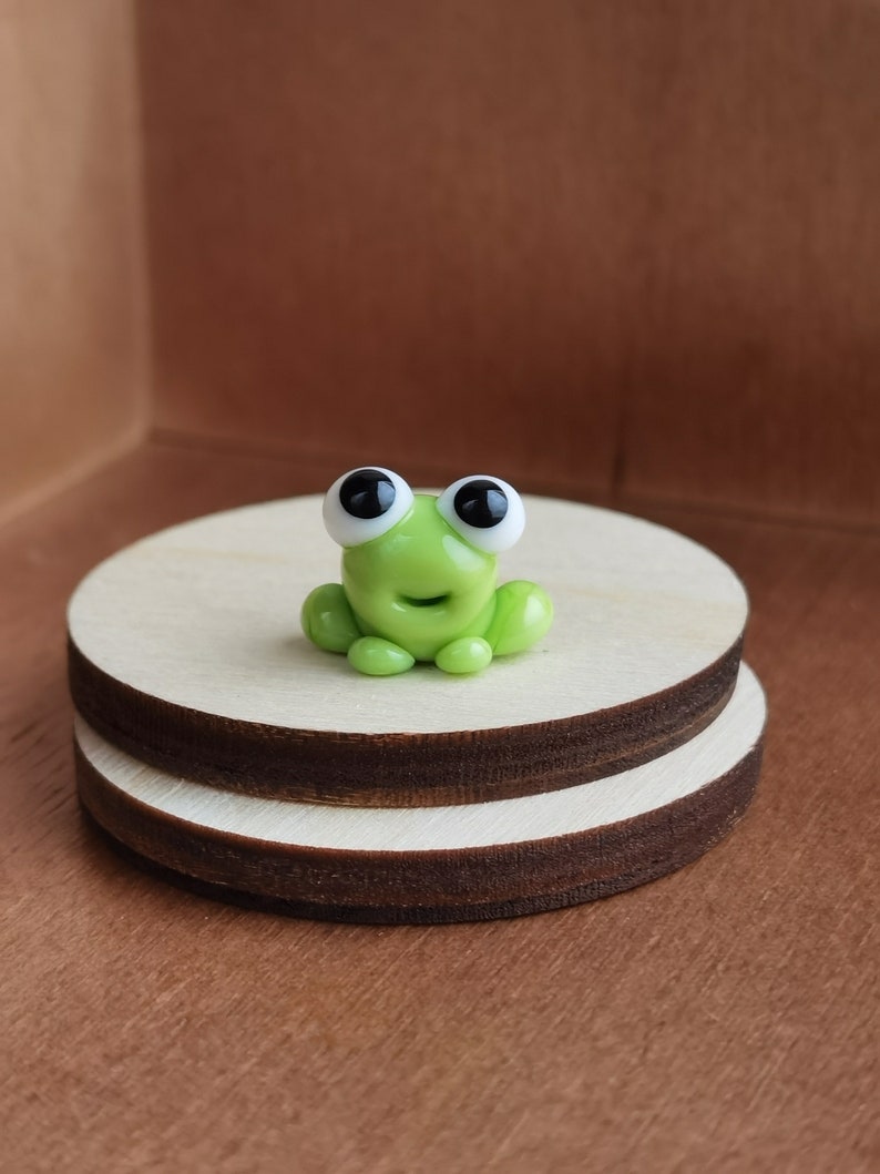May include: A small green glass frog figurine with black eyes and white pupils. The frog is sitting on a wooden platform.