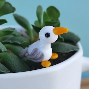 May include: A small, handcrafted glass duck figurine with a white body, gray wings, and orange beak and feet. The duck is set in a white ceramic pot with green succulent plants, against a light blue background. A charming decorative item.