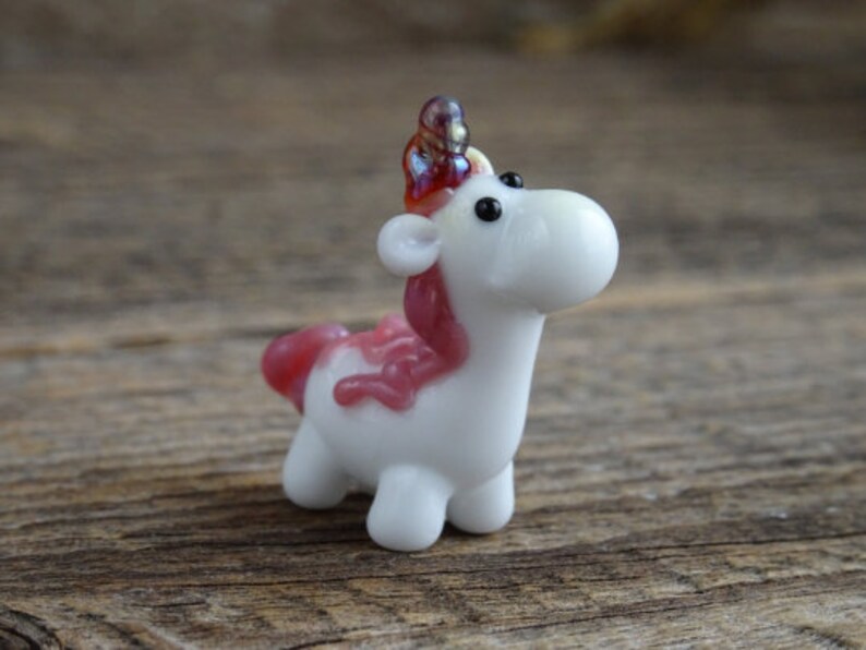 Op de afbeelding: Een klein wit glazen eenhoornbeeldje met een roze manen en hoorn. De eenhoorn heeft zwarte ogen en een wit lichaam.