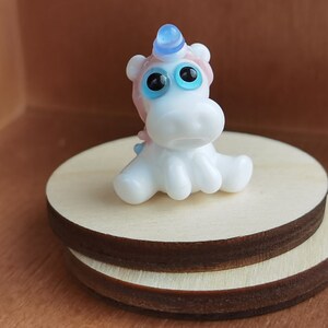 May include: A small white glass unicorn figurine with blue eyes and a blue and pink horn. The unicorn is sitting on a wooden base.