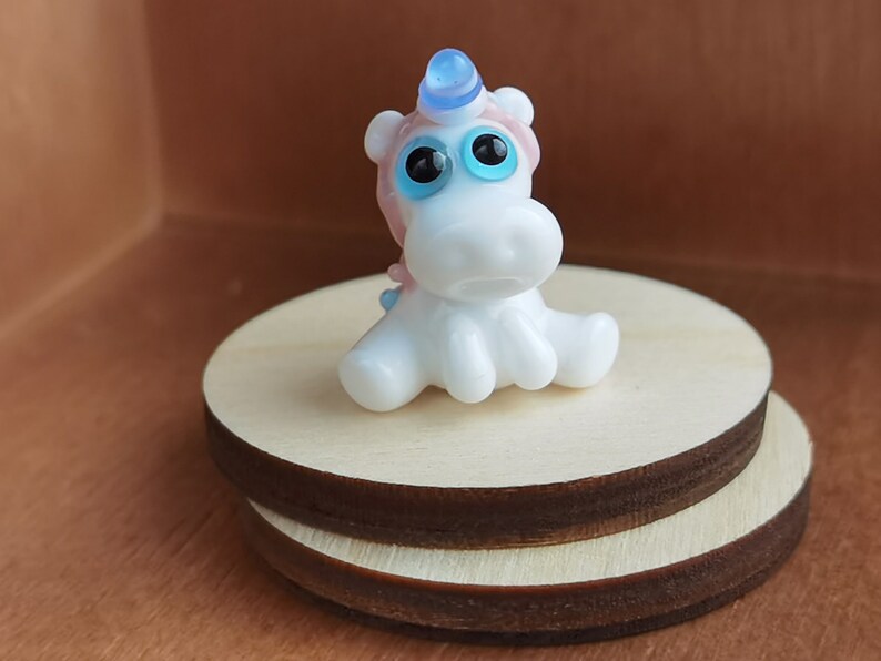 May include: A small white glass unicorn figurine with blue eyes and a blue and pink horn. The unicorn is sitting on a wooden base.