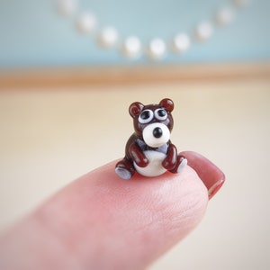 May include: A small, brown glass bear figurine with white eyes and a white belly. The bear is sitting on a finger.