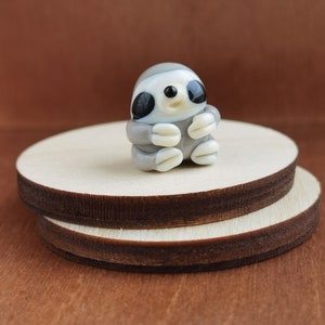 May include: A small glass sloth figurine with a light brown body, white belly, and black eyes. The sloth is sitting with its arms crossed and has a small smile on its face.