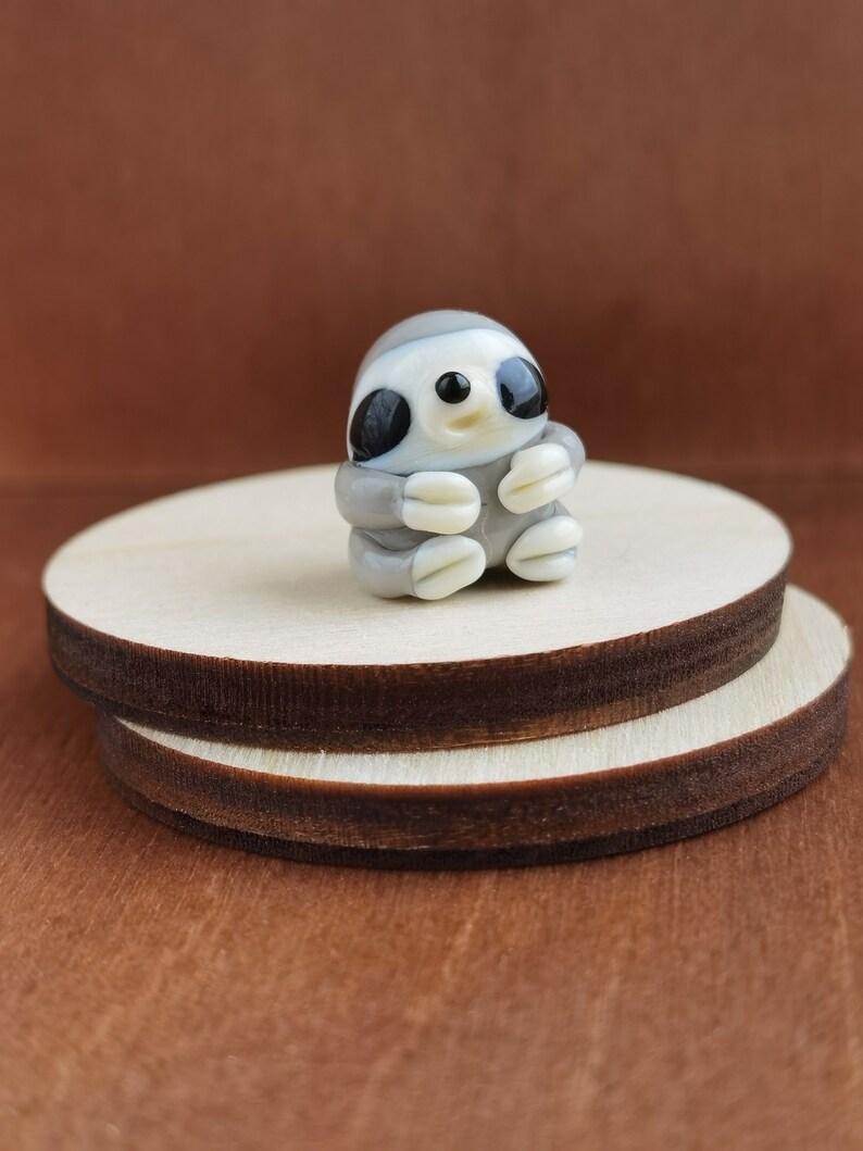 May include: A small glass sloth figurine with a light brown body, white belly, and black eyes. The sloth is sitting with its arms crossed and has a small smile on its face.