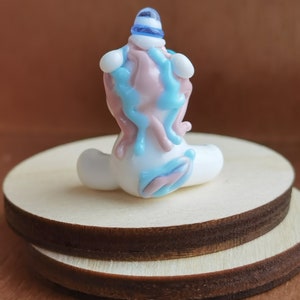 May include: A small glass figurine of a unicorn with a blue and white hat. The unicorn is sitting on a wooden base and is made of white, pink, and blue glass.