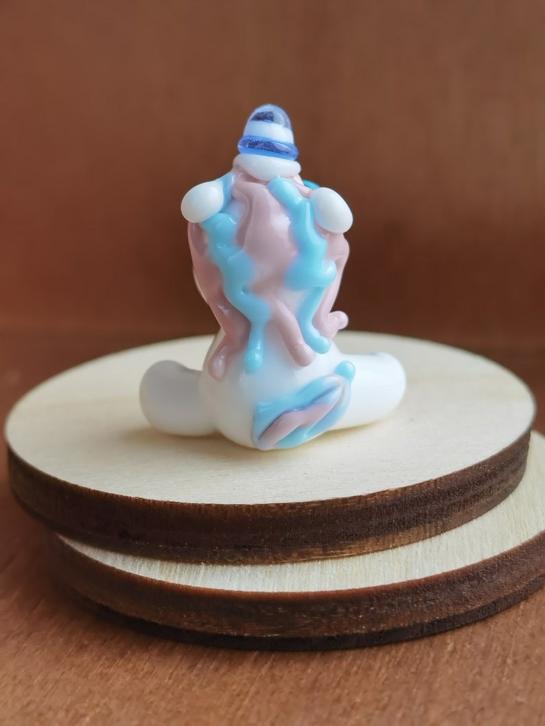 May include: A small glass figurine of a unicorn with a blue and white hat. The unicorn is sitting on a wooden base and is made of white, pink, and blue glass.