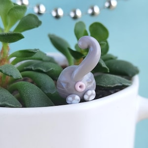 May include: A small gray ceramic figurine of a cat's rear end with a pink tail and white paw prints. The figurine is sitting in a white ceramic pot with green succulent plants.